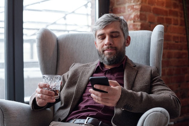 Contenido empresario canoso con barba sentado con vaso de whisky y desplazamiento en smartphone