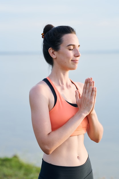 Contenido hermosa chica en sujetador deportivo manteniendo las manos en Namaste mientras agradece en cuerpo y alma después de la práctica de yoga