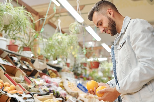 Contenido joven barbudo en chaqueta de mezclilla de pie en el mostrador de alimentos y elegir entre dos naranjas