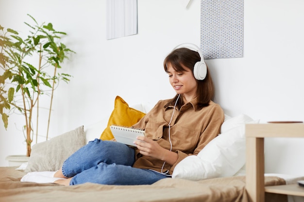 Contenido joven creativa en auriculares blancos sentado en la cama con cómodas almohadas y haciendo n