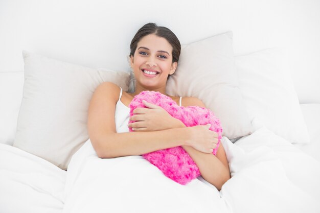 Contenido joven modelo de cabello castaño en pijama blanco abrazando una almohada en forma de corazón