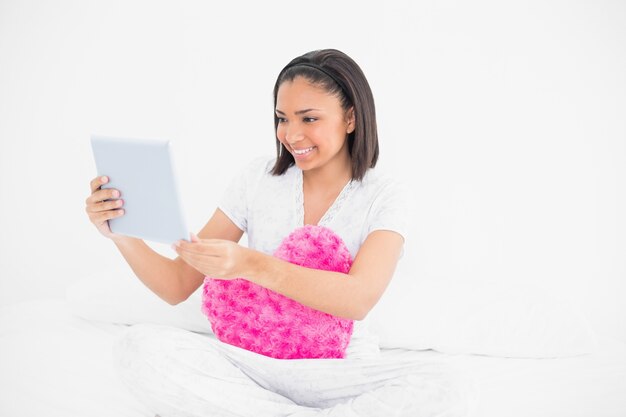 Contenido joven modelo de pelo oscuro abrazando una almohada y usando una tablet pc