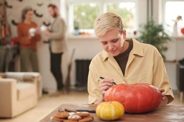 Contenido joven rubia con camisa amarilla sentada en la mesa y dibujando el diseño de la cara en la calabaza mientras