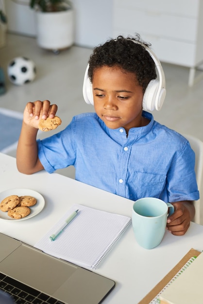 Contenido un lindo niño afroamericano con auriculares bebiendo té con galletas y viendo videos en línea en una computadora portátil