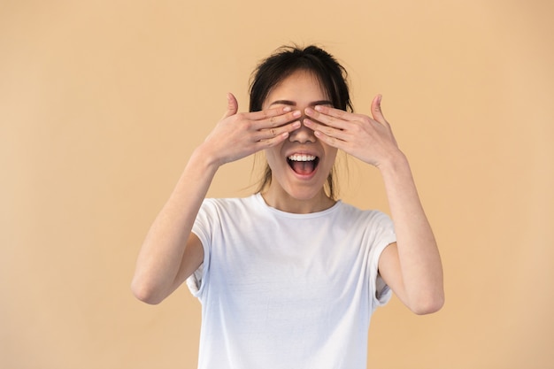 Contenido mujer china vistiendo camiseta básica cubriéndose los ojos con las manos aisladas sobre pared beige en studio