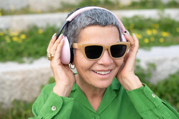 Contenido senior mujer escuchando música en el parque. Modelo femenino con cabello gris corto en ropa brillante y grandes auriculares. Ocio, actividad, concepto de tecnología.