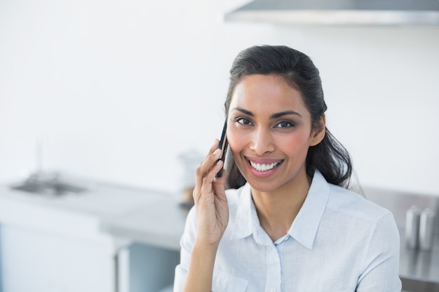 Contenido sonriente mujer llamando con su teléfono inteligente en la cocina brillante