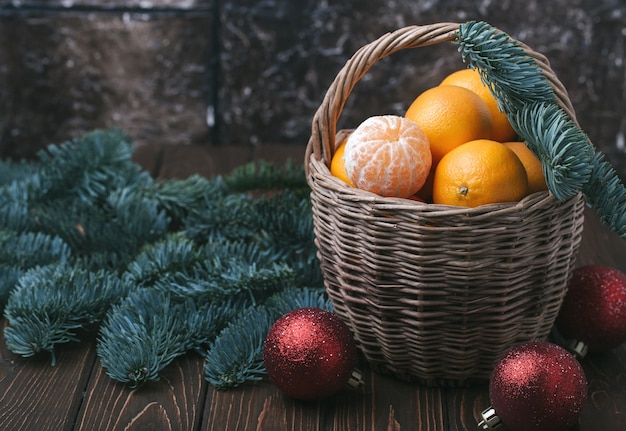 Foto conteúdo de férias, tangerinas, tangerina descascada em uma cesta de vime, vintage, galho de abeto, bolas vermelhas de árvore de natal, fundo marrom escuro, horizontal