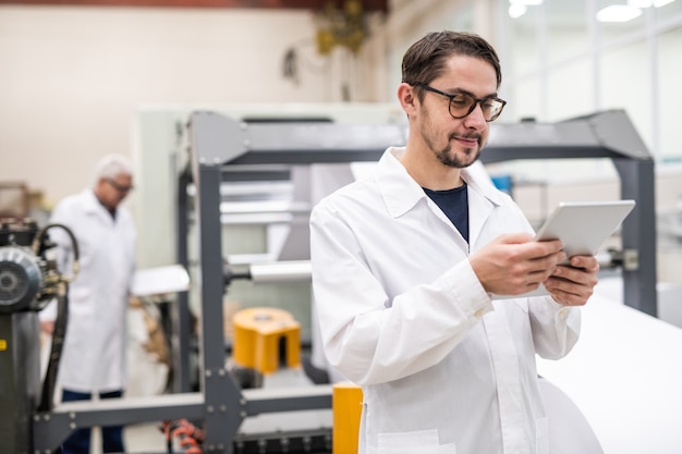 Conteúdo jovem barbudo especialista em óculos usando tablet digital enquanto analisa recursos de impressão na fábrica