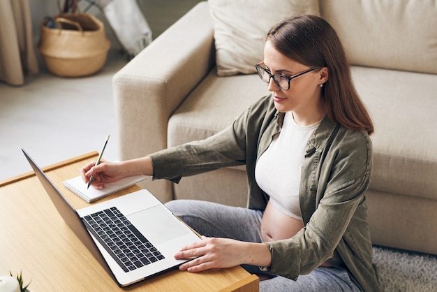 Conteúdo, jovem grávida, usando óculos, sentada à mesa de centro e usando um laptop enquanto faz anotações sobre a maternidade em casa