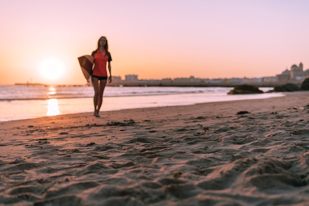 En el contexto de una magnífica puesta de sol en el océano, una niña camina con una tabla de surf bajo el brazo. Se puede ver una gran ciudad en la distancia.