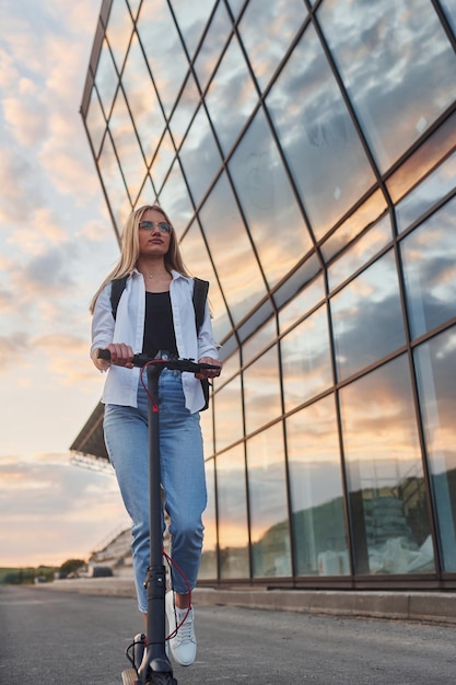 Contra a construção de negócios modernos Linda loira em roupas casuais andando de scooter elétrica ao ar livre no dia ensolarado