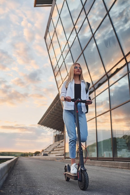 Contra a construção de negócios modernos Linda loira em roupas casuais andando de scooter elétrica ao ar livre no dia ensolarado