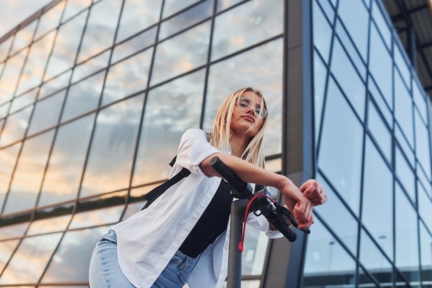 Contra a construção de negócios modernos Linda loira em roupas casuais andando de scooter elétrica ao ar livre no dia ensolarado