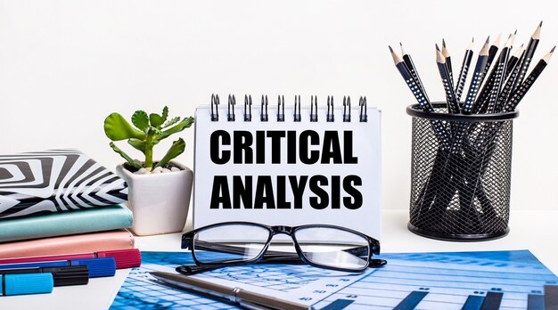 Contra a parede de um esquema azul e uma parede branca, lápis pretos em um suporte, uma flor, diários e um caderno com a inscrição ANÁLISE CRÍTICA