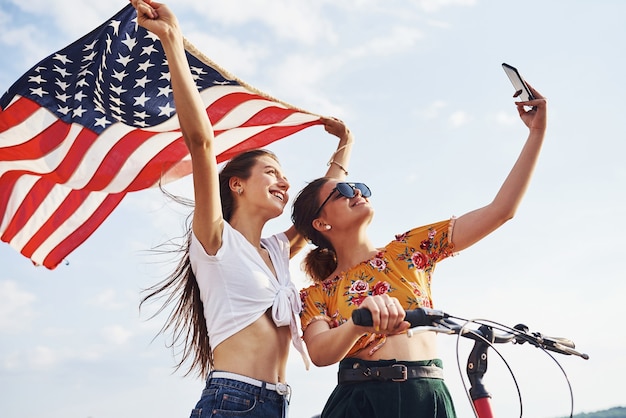 Contra el cielo nublado. Dos mujer alegre patriótica con bicicleta y bandera de Estados Unidos en manos hace selfie.