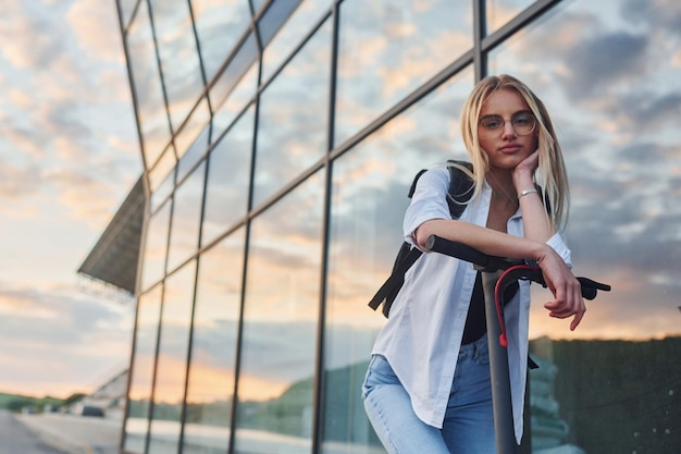 Contra el edificio de negocios moderno Hermosa rubia en ropa casual montando scooter eléctrico al aire libre en el día soleado