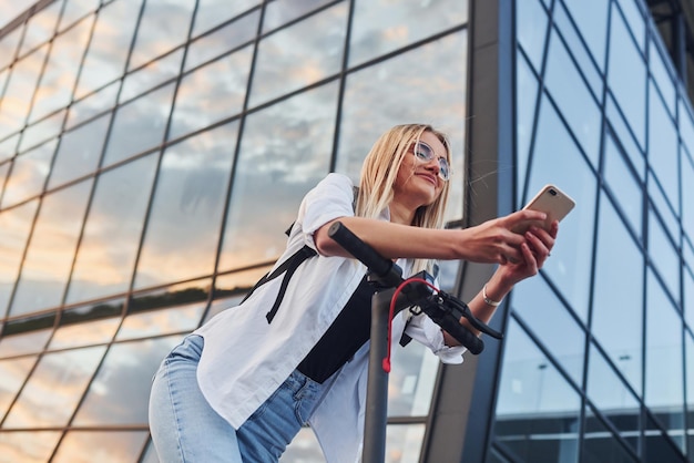 Foto contra el edificio de negocios moderno hermosa rubia en ropa casual montando scooter eléctrico al aire libre en el día soleado