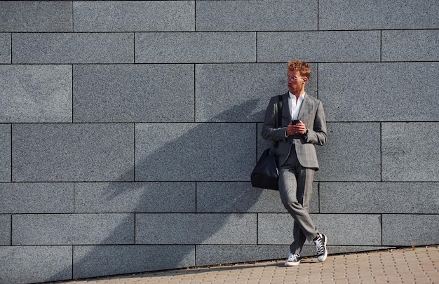 Contra la pared gris Joven empresario en ropa formal gris está al aire libre en la ciudad