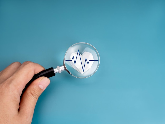 Control médico del corazón concepto de atención médica Línea de medición del latido del pulso en fuerte enfoque cardíaco blanco geométrico en lente de lupa sostenida por la mano humana aislada en fondo azul minimalista