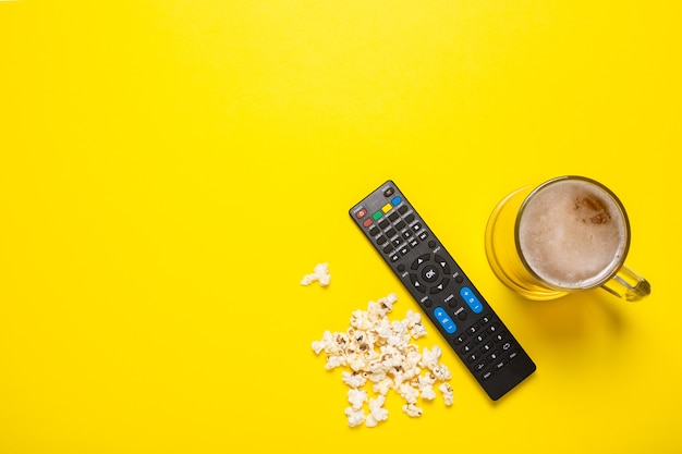 Control remoto de un televisor o sintonizador de TV, un vaso de cerveza y palomitas de maíz en amarillo