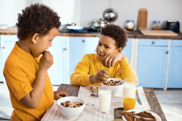 Conversa agradável. Garotos pré-adolescentes animados sentados à mesa e conversando enquanto comem cereais no café da manhã