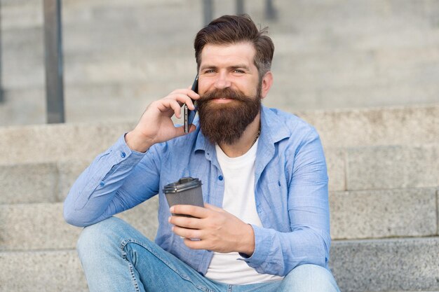 Conversa fiada com alguém Hipster feliz bebe café falando ao telefone ao ar livre Fazendo uma ligação telefônica Conexão de celular Nova tecnologia Vida moderna O celular facilita a comunicação