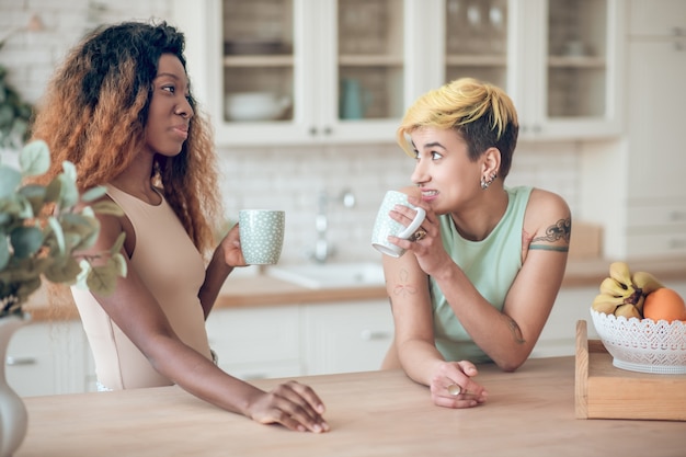 Conversa interessante. Mulher amaricana de cabelos compridos sorridente e namorada caucasiana surpresa com cabelo curto tingido tomando café na cozinha