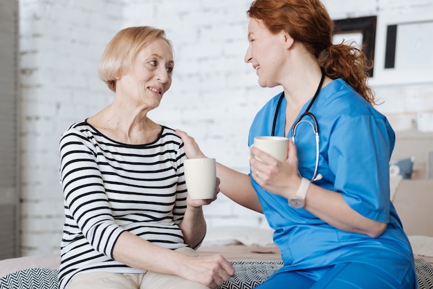 Conversação como terapia. Admirável bela senhora positiva oferecendo uma xícara de chá à jovem como sinal de gratidão por ajudá-la a se recuperar de uma doença