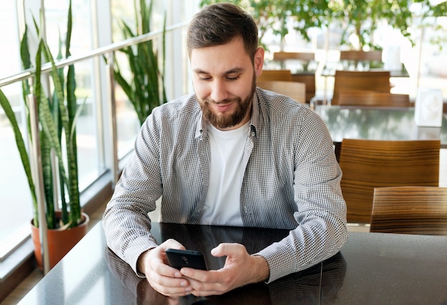 Conversación telefónica. Retrato sonriente hombre de barba inteligente en la oficina moderna con teléfono inteligente, mensajes de texto en la aplicación, consultar el correo electrónico, navegar por la web de internet. Empresario atractivo independiente en café