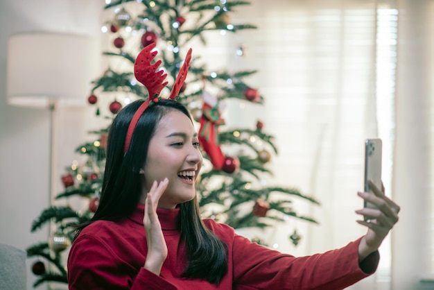 Conversación de videollamada de mujer asiática atractiva a través de Smartphone Saludo Navidad Año Nuevo Fondo de árbol de Navidad