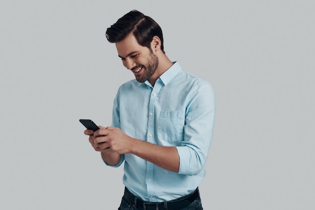 Converse com um amigo. jovem bonito usando um telefone inteligente e sorrindo em pé contra um fundo cinza