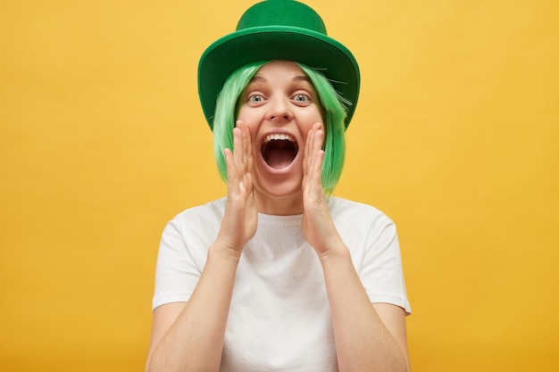 Convidando para a festa do dia de São Patrício Uma mulher caucasiana excitada vestindo um chapéu verde de duende de pé isolada sobre um fundo amarelo gritando com as mãos perto da boca fazendo um anúncio