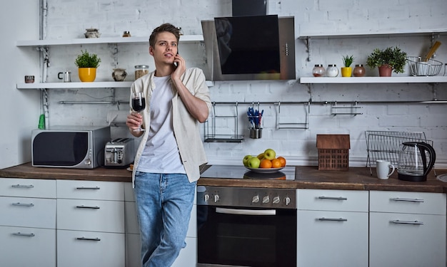 Convidar amigos para jantar. Jovem bonito com roupas casuais bebendo vinho e falando ao telefone enquanto fica sentado na cozinha de casa