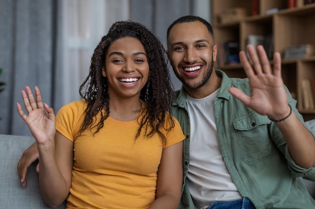 Cónyuges afroamericanos alegres saludando con la mano y sonriendo a la cámara hablando en línea a través de una videollamada