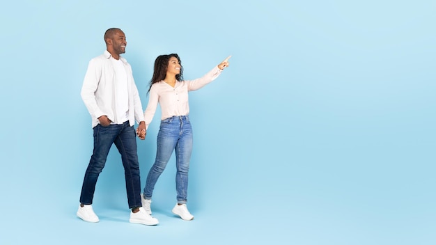 Cónyuges afroamericanos felices caminando y tomados de la mano mujer señalando con el dedo el panorama de fondo azul del espacio libre