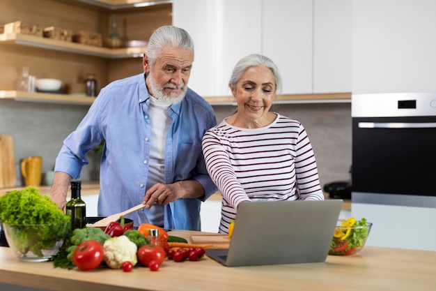 Cónyuges ancianos alegres que miran la receta en la computadora portátil mientras que cocinan la comida en cocina