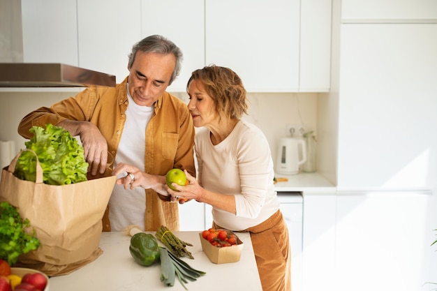 Cónyuges ancianos desempacando bolsas de papel con verduras y frutas en la cocina pidiendo comida o viniendo