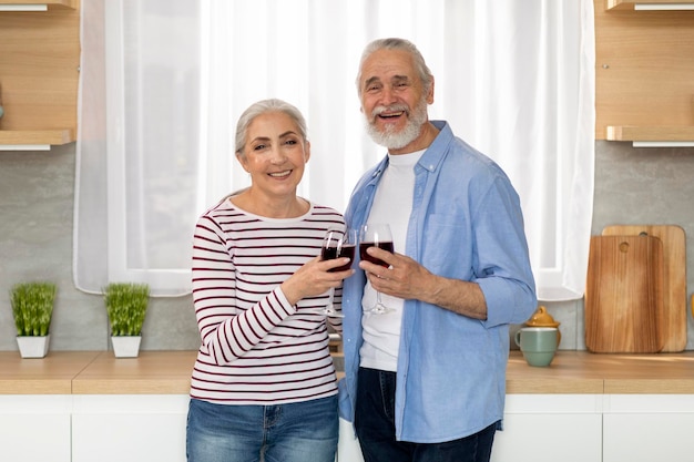 Cónyuges ancianos sonrientes sosteniendo copas con vino mientras posan en el interior de la cocina