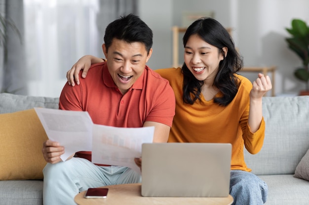 Cónyuges asiáticos emocionales leyendo documentos usando una computadora portátil