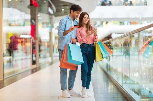 Cónyuges felices comprando con teléfono inteligente y tarjeta de crédito en el centro comercial