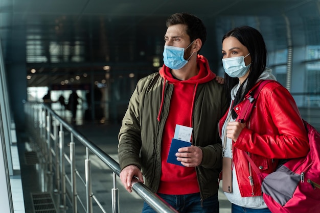 Cónyuges felices con máscaras protectoras paradas frente a la ventana y mirándola en la sala del aeropuerto. Viajar durante el concepto de pandemia