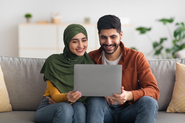 Cónyuges felices del medio oriente usando una computadora portátil sentada en el sofá interior