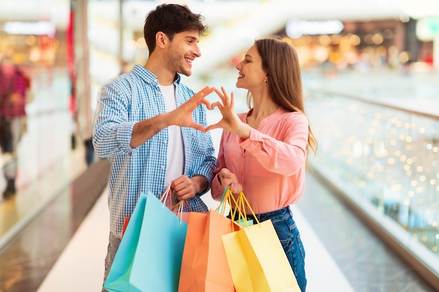 Foto cónyuges haciendo dedos corazón sosteniendo bolsas de compras en el centro comercial