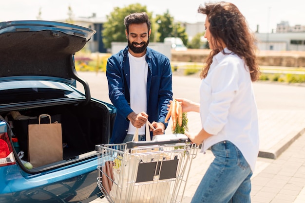 Cónyuges jóvenes del Medio Oriente empacando comestibles en el baúl del auto al aire libre de pie en el estacionamiento cerca