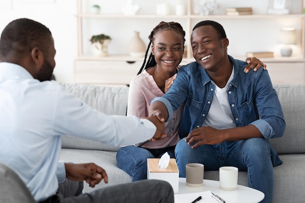 Cónyuges negros felices agradecidos con el consejero familiar después de una exitosa sesión de terapia marital, estrechando la mano con el psicoterapeuta, agradecidos por la reconciliación, sentados en el sofá en la oficina, espacio libre