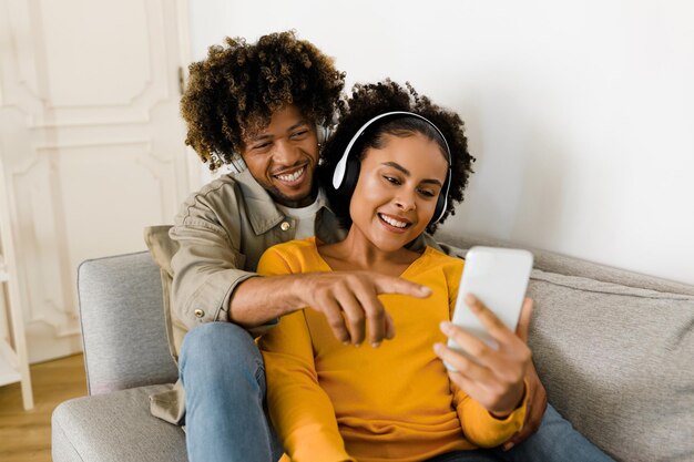 Cónyuges negros felices usando el teléfono escuchando música juntos en el interior