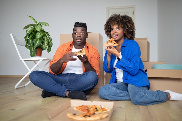 Cónyuges negros jóvenes descansando en el día de la mudanza comiendo pizza y viendo la televisión