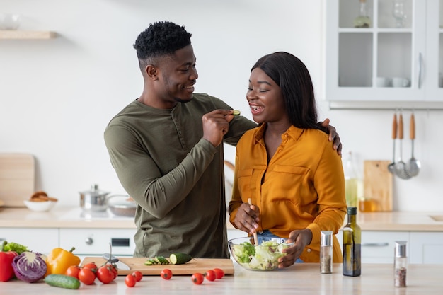 Cónyuges negros románticos divirtiéndose mientras preparan comida en una cocina acogedora