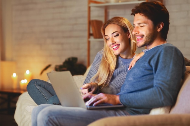 Cónyuges sonrientes usando la computadora portátil sentados en el sofá en casa